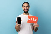 Man wearing white T-shirt holding sale card and smartphone, being satisfied with online sell-out.