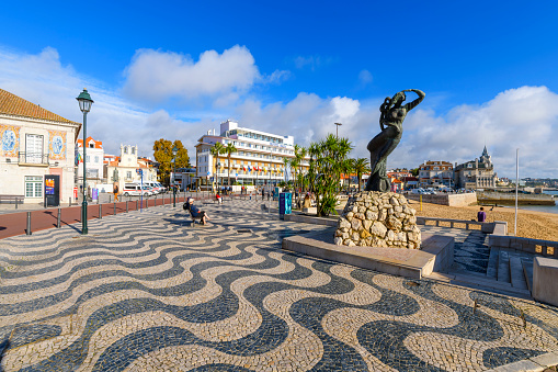 A wide sidewalk of calçada portuguesa, the traditional-style patterned pavement used for many pedestrian areas in the historic seaside old town of the resort town of Cascais, Portugal, with the sandy beach, city hall and Palacete Seixas in view behind.