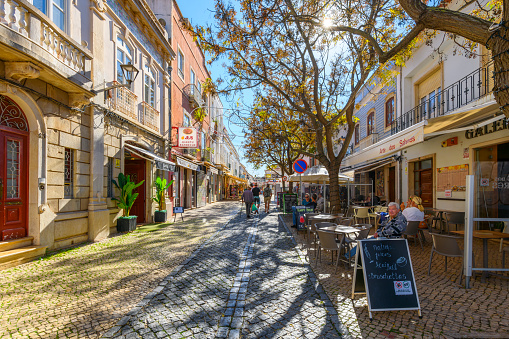 Shady treed pedestrian cobblestone street of shops and sidewalk cafes in the historic old town center of the seaside village of Lagos, Portugal at autumn. Lagos is a town in southern Portugal's Algarve region. It’s known for its walled old town, cliffs and Atlantic beaches.