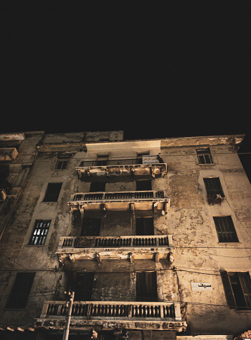 This is a photo of an old and dilapidated building that was once a luxurious mansion. The building has three floors and balconies with ornate railings. The photo is taken at night, and the building is illuminated by streetlights. The building has a mysterious and eerie atmosphere, as if it hides secrets from the past.