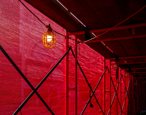 Close-up of a construction scaffolding frame and wood planks over a sidewalk. The scaffolding is covered in red netting and is illuminated by a single yellow light fixture and bulb.