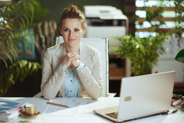 Portrait of woman worker in light business suit in green office Portrait of modern middle aged woman worker in a light business suit in modern green office with laptop. sales executive stock pictures, royalty-free photos & images