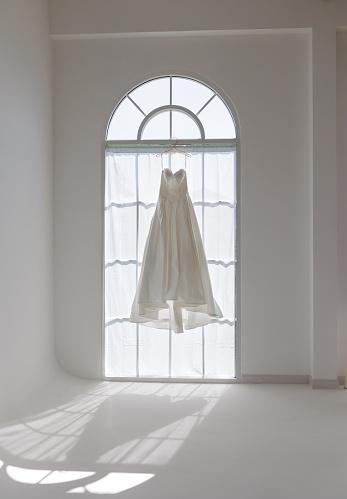 The gentle morning sun filters through a sheer curtain, casting a radiant glow on a solitary wedding dress hanging before a vintage arched window, awaiting the day's joyous celebrations