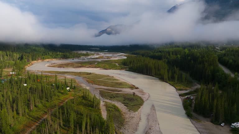 Drone shot pushing forward and tilting up to reveal mountains with low hanging clouds over the Kicking Horse River in British Columbia Canada. People have camp sites set up on the riverbanks.