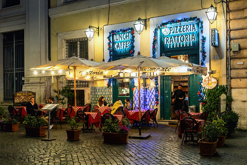 Rome, Italy - January 05, 2023: People sitting in a small outdoor restaurant on cobblestone street illuminated in the evening in Rome - capital and the most populous city in Italy, famous and popular travel destination.