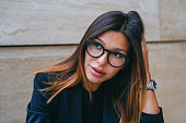 Close up portrait of brunette young businesswoman in black suit, spectacles, looks away with pensive face expression sitting outside. Puzzled american woman in thoughtful mood.