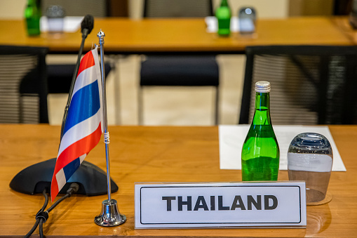 Name plate holder sign with Thailand flag, gooseneck paging microphone, notes and water on a table in conference hall during summit, meeting, forum or other event