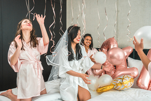 Diverse bridesmaids and a joyous Hispanic bride, all in silk bathrobes, enjoy playful moments with balloons in the hotel room during bachelorette party preparations.