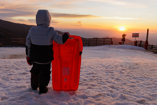 Boy with sled standing backlit at sunset