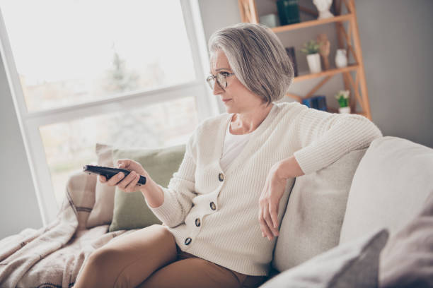 かわいい肯定的な年配の女性が白いカーディガンのアイウェアを着てソファに座って屋内の部屋の部屋で映画を楽しんでいる写真 - senior women electronics people remote control ストックフォトと画像
