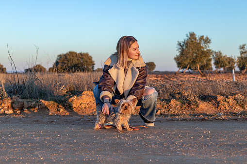 Mota del Cuervo, Castilla La Mancha, Spain. A young woman in a squatting position, caressing her little dog while gazing into the distance in a rural setting at sunset.