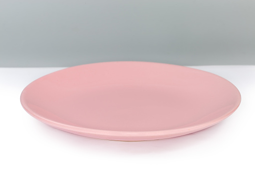 Empty glazed ceramic pink shallow plate for serving food and dining on a white surface, side view on a gray background