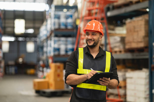istock Professional manager man employee smile using tablet check stock working at warehouse. Worker wearing high visibility clothing and a hard hat, helmet and checking and count up goods boxes delivery. 1826553972