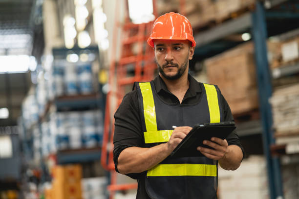 istock Professional manager man employee using tablet check stock working at warehouse. Worker wearing high visibility clothing and a hard hat, helmet and checking and count up goods or boxes for delivery. 1826553973