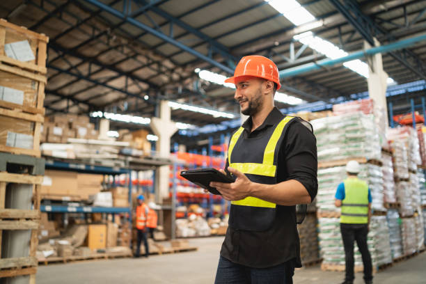 istock Professional manager man employee smile using tablet check stock working at warehouse. Worker wearing high visibility clothing and a hard hat, helmet and checking and count up goods boxes delivery. 1826554098
