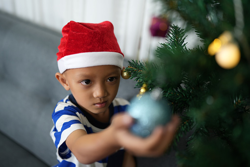 Merry Christmas 2024 concept Asian boy decorates Christmas tree with colorful Christmas balls.