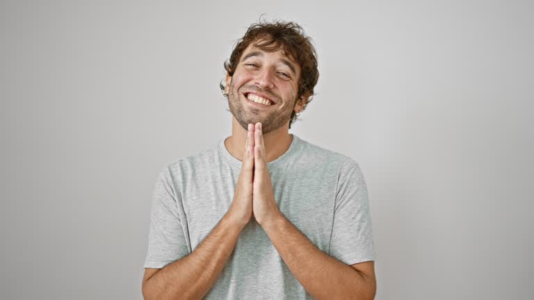 Confident young man in casual t-shirt, praying with hands together, asking god for forgiveness on isolated white background, hopeful smile lighting his face