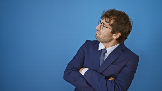 Portrait of a confident young man in business suit, arms crossed, looking to the side with a cheerful look on isolated blue background
