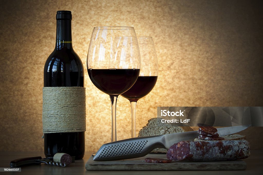 Bottle of wine, glasses, salami and bread. "Original hand-made wine bottle with two glass of red wine and some slices of bread, salami, and contemporary kitchen knifeNice windows reflections and warm colors.Backlit effect." Alcohol - Drink Stock Photo