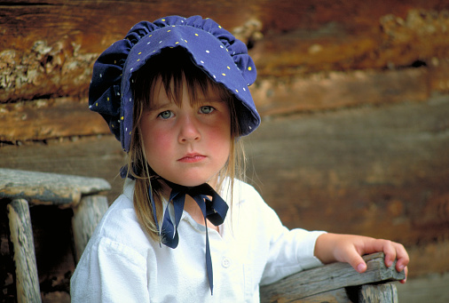 Serious-looking Pioneer girl with bonnet sitting on  the porch of a log cabin.