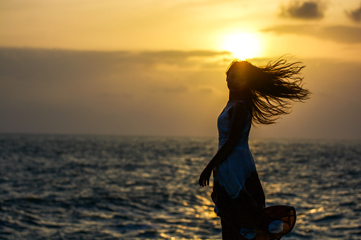 Asian Young woman enjoying freedom at the beach. Beautiful female model under sunset at seaside. Calm water reflects woman silhouette. Sun goes behind horizon. Girl is alone.