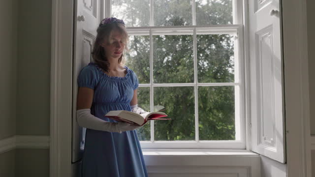 Young woman wearing a regency era dress is reading a book near a window