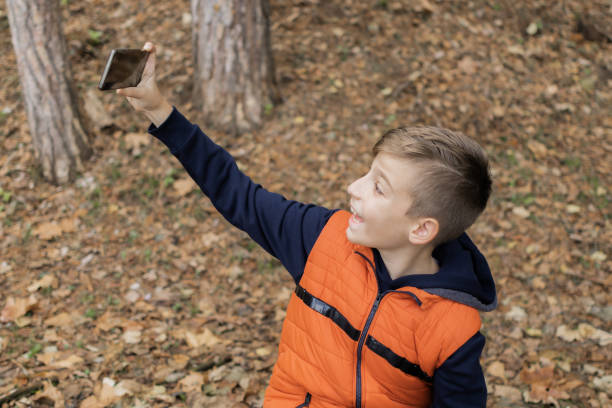 istock Cute schoolboy is taking selfie or making a video call with his mobile phone. Boy is sitting on the bench in public park in autumn. He is happy. 1826652115