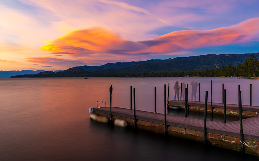 This August 2020 long-exposure image shows the waters of Lake Te Anau in Aotearoa New Zealand at sunrise on a cloudy morning. A few spots of the morning sun shine through the clouds leaving a warm glow. Smooth, multi-coloured stones are seen on the lakeshore.