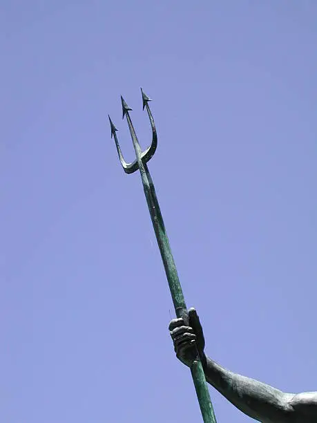 Part of a statue with an arm holding a trident with blue sky in the background