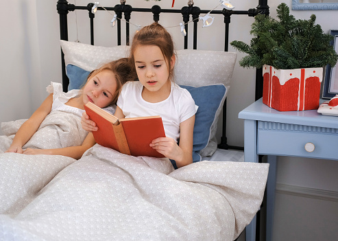 Two little sisters are reading a book in bed while in a room decorated with Christmas decorations and garlands