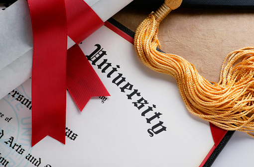 A red ribbon wrapped around a rolled up diploma sits on top of a another college diploma.  A gold tassel from a graduation cap is also draped across the diploma.
