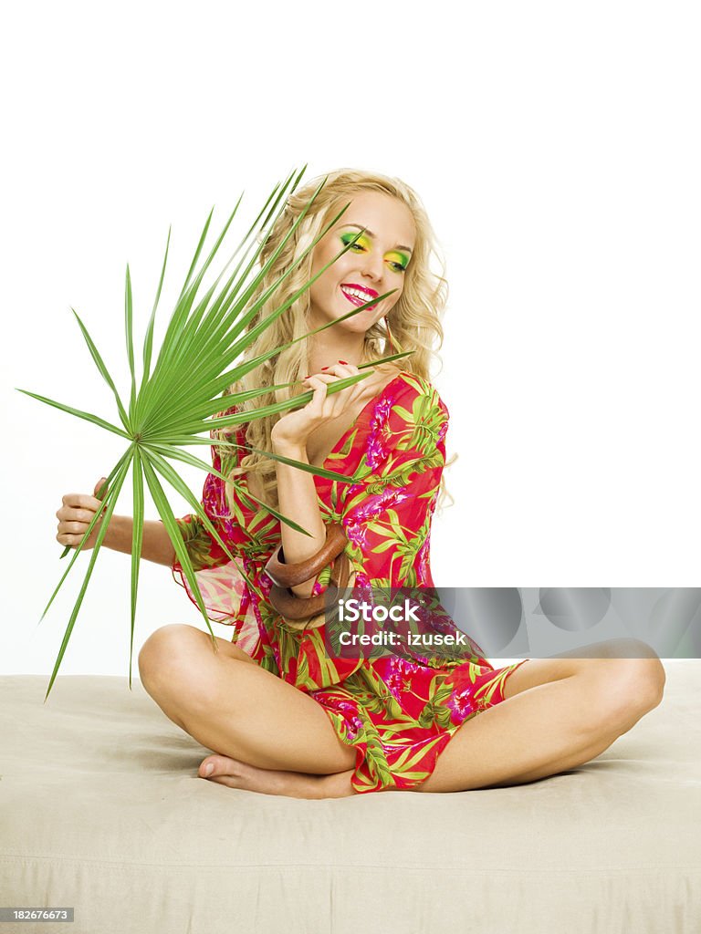 Beautiful blonde woman holding large tropical leaf. "Summer portrait of a beautiful blonde woman sitting with her legs crossed,wearing a colorful tunic. Holding large tropical leaf." 20-24 Years Stock Photo