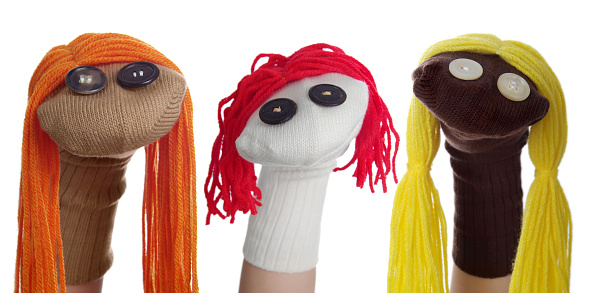 Horizontal image of three sock puppets lined in a row on a white background. One puppet was created using a tan sock with orange hair, one puppet was made with a white sock and red hair, the third puppet was created with a dark brown sock with yellow hair. Their heads are tilted different directions giving them different expressions.