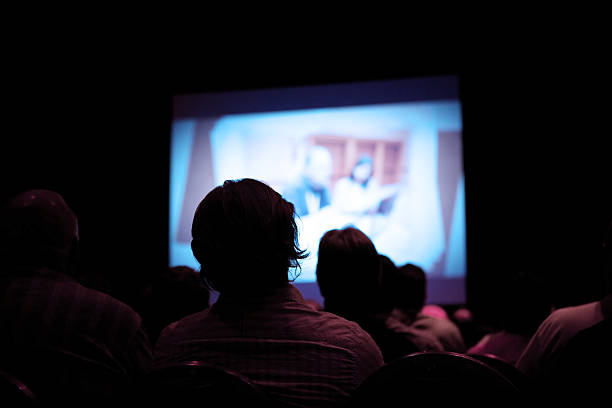 People watching movie in dark cinema An unknown number of people sitting in a dark theater.  There is a projector screen in front of them with a man and woman at a conference. projection screen stock pictures, royalty-free photos & images