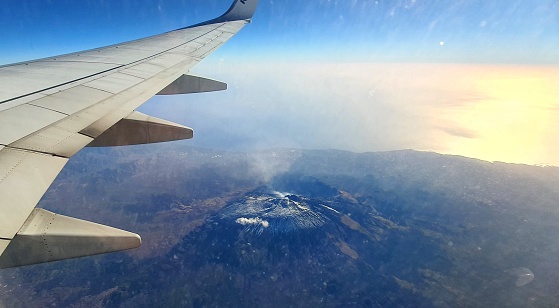 Flying from Malta in the November morning. Aerial view on Etna volcano - highest European active stratovolcano. Snow and smoke visible on the mountain. Wing of an aircraft in the picture.