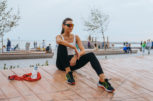 strong tanned runner is resting after a marathon in the park dressed in sports clothes sneakers sunglasses. Young woman training outdoors. Women sports.