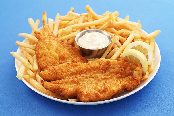 Full-frame, high-resolution digital capture of a plate of delicious, crispy fried fish and chips with a metal ramekin of tartar sauce. Plate sits on a blue surface, and is garnished with lemon slices.