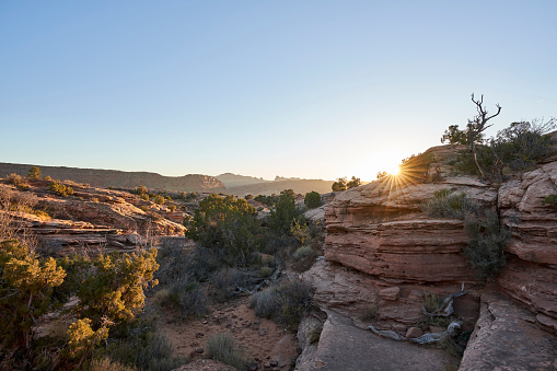 The evening sun goes down over the rocky desert. The rocky and sandy terrain is composed of rock formations and sparse grass and shrubs.