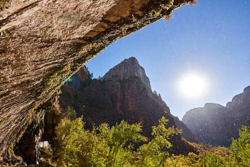 Water drips from a rock overhang. The low sun shines up the canyon backlighting the fall trees and water droplets. The valley is surrounded by rock peaks.