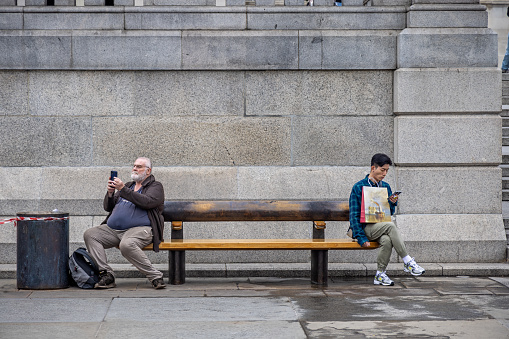 Trafalgar Square, London, England - October 25th 2023: Two men on a bench - one mature European and one young Asian with their mobile phones