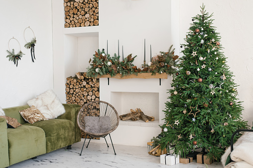 Rustic home interior of the living room with a Christmas wreath, fireplace, firewood, sofa and Christmas tree. Decorations for the celebration of Christmas holidays