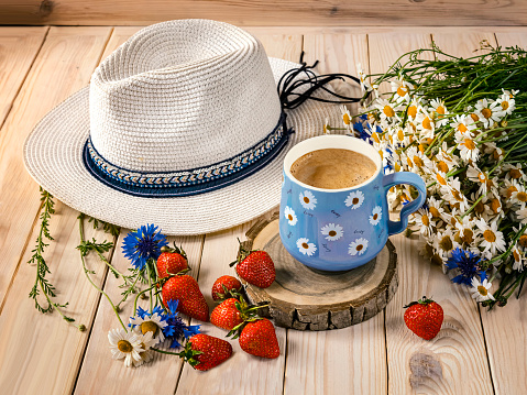 A blue cup of coffee with daisies, a bouquet of daisies and cornflowers, strawberries, a hat from the sun. Summer still life with wildflowers, top view