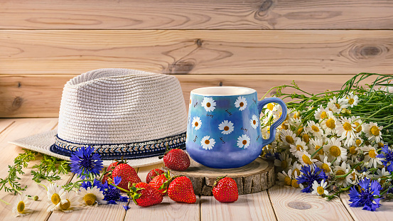 A blue cup of coffee with daisies, a bouquet of daisies and cornflowers, strawberries, a hat from the sun. Summer still life with wild flowers.