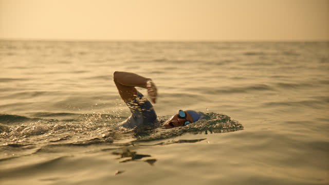 SLO MO Lockdown shot of determined mid adult woman swimming in seascape against sky during sunset. Female swimmer swimming front crawl at sea in sunshine