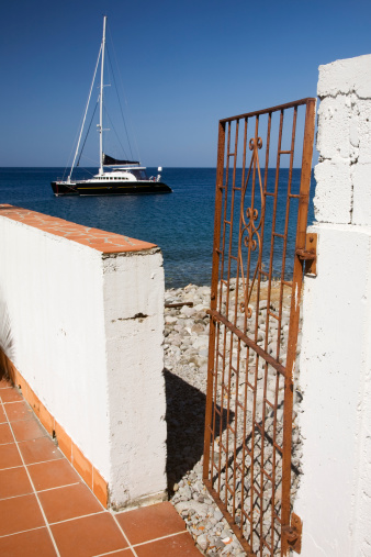 A rusty metal gate is open to the rocky beach that leads to the Caribbean sea. A catamaran sailboat sits in the background. Taken in Dominica. See my other images from Dominica: