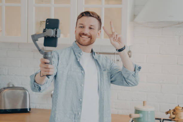 istock Cheerful man making a peace sign while filming with a smartphone on a gimbal in a kitchen 1827042145