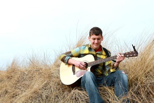 A 17 year old boy smiles as he plays his acoustic guitar outside at the beach. Plenty of copy space in sky.