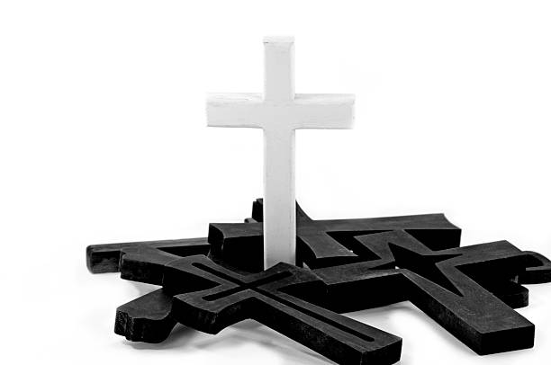 A white rough white wood cross standing among several black fallen crosses. White background in black and white with copy space.