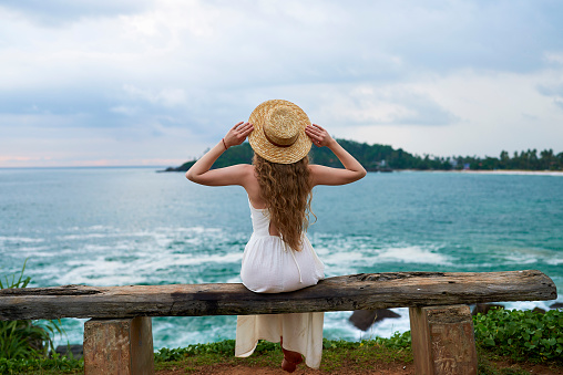 Young woman in white dress sits on rustic bench by the sea, contemplating waves. Back view of female in straw hat enjoys ocean landscape, solitude moment, tropical coast.