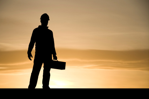 A construction worker stands with a toolbox in hand against a rising sun.